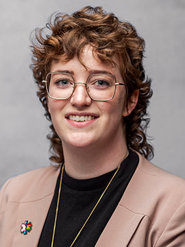 a professional headshot of a white nonbinary person with curly hair and glasses, wearing a LGBT pride flag pin.